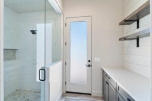 Custom home guest bathroom featuring a glass-enclosed shower, dark vanity with white countertops, and a frosted glass door providing natural light and private outdoor access in Arizona