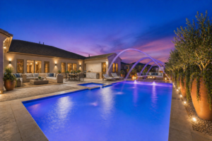 A luxury San Tan Valley backyard featuring a brightly lit blue swimming pool with arched laminar water jets, a full outdoor kitchen, and a lounge area with an outdoor fireplace at twilight.