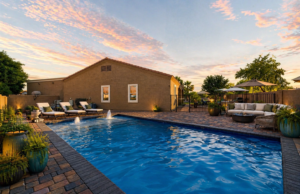 Integrated design-build backyard in Queen Creek, AZ, featuring a large geometric swimming pool with fountain bubblers, stone paver decking, and a modern lounge area at sunset.