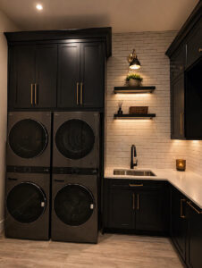 Modern custom laundry room featuring black stacked washers and dryers, dark cabinetry, and illuminated floating shelves.